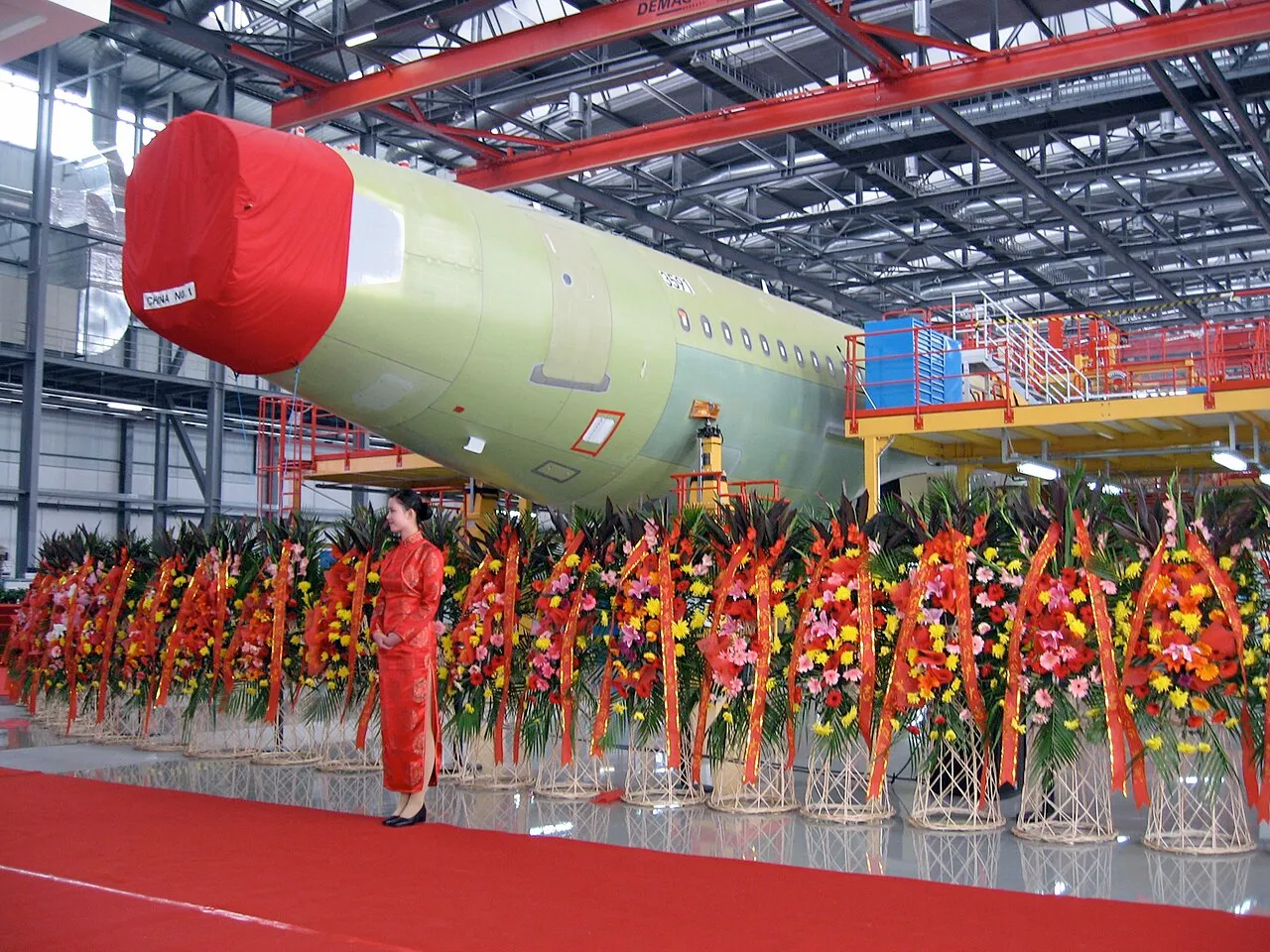 Aircraft fuselage inside an assembly hall with work platforms and floral displays.
