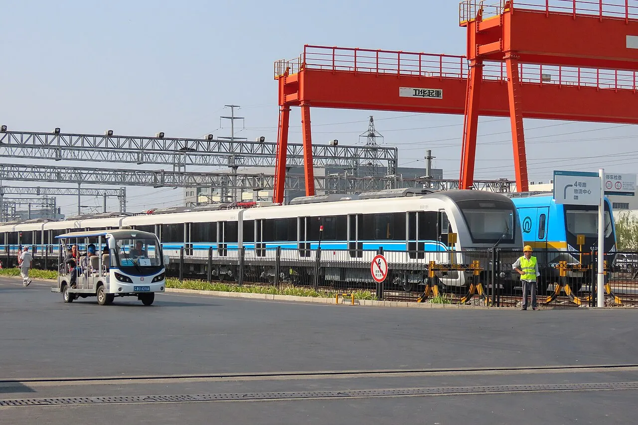 Metro trains beneath red gantry cranes on a factory site.